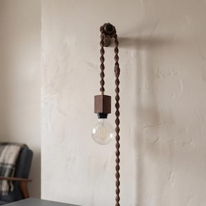 May include: A modern wall-mounted light fixture with a brown macrame cord. The fixture features a wooden square base and a clear, round lightbulb. The cord is attached to a wooden decorative element on the wall. The light is hanging against a neutral-colored wall.