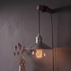 Op de afbeelding: Een hanglamp in vintage-stijl met een geborstelde metalen kap en een grote, zichtbare gloeidraadlamp. De lamp hangt aan een houten balk die aan een donkergrijze muur is bevestigd. Het zwarte snoer is om de balk gewikkeld.
