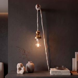 May include: A wall-mounted pendant light featuring a brass fixture with a spherical and dome-shaped top, suspended by a white twisted fabric cord knotted around a wooden peg. A clear glass globe bulb with visible filaments hangs from the fixture.