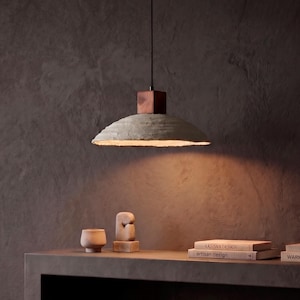 May include: A pendant light featuring a wide, textured, light gray concrete shade suspended by a thin black cord from a dark brown wooden block. The light emits a warm, downward glow, illuminating the surface below with a soft, focused light.