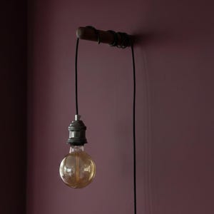 May include: A wall-mounted light fitting with a wooden support and a black cable. The fitting has a vintage-style Edison bulb with a warm, amber glow. The cable is wrapped around the wooden support. The background is a solid, dark pink.