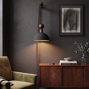 May include: A contemporary, dark grey pendant lamp with a dome-shaped shade suspended from a brown rope and wooden fitting. The lamp casts a warm light downwards. It's mounted on a dark grey wall, with a wooden cabinet and framed artwork in the background.