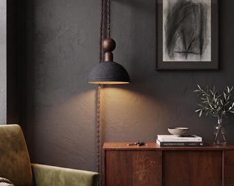 May include: A modern, dark gray pendant lamp with a dome-shaped shade hangs from a brown rope and wooden fixture. The lamp illuminates a warm glow downwards. The lamp is mounted on a dark gray wall, with a wooden cabinet and a framed artwork in the background.