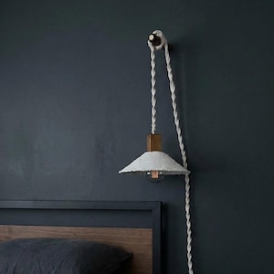May include: A white paper shade pendant light with a wooden block and a twisted rope cord. The light hangs from a wooden peg on a dark blue wall. The light fixture is above a bed with a dark wooden headboard.