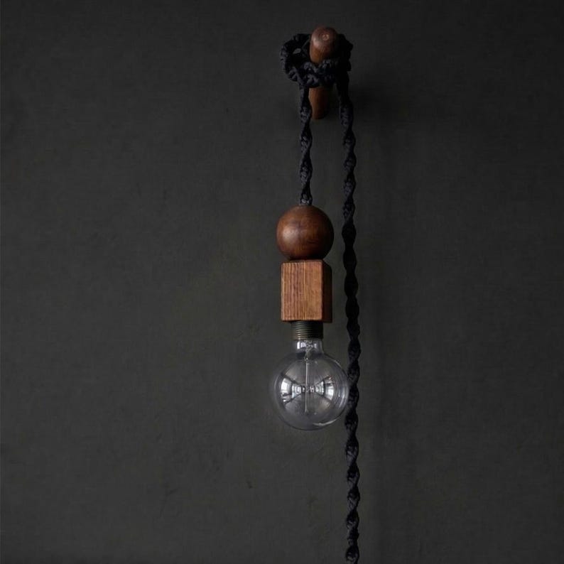 Peut inclure: Suspension luminaire moderne avec une ampoule en verre transparent, une sph&egrave;re et un bloc carr&eacute;s en bois. Le luminaire est suspendu par un cordon torsad&eacute; noir et mont&eacute; sur un mur sombre. Les &eacute;l&eacute;ments en bois ont une finition naturelle et chaleureuse.