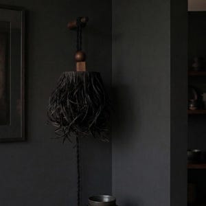 以下が含まれることがあります： 暗い壁に木製の器具から吊り下げられた、黒いフリンジ付きのランプシェード。 ランプシェードは、密度の高い羽のような質感の円筒形をしています。 暗いコードが下方に伸び、小さなボウルが下にあります。