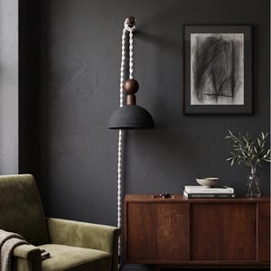 May include: A contemporary pendant light featuring a black lampshade and wooden detailing. It is suspended from a white rope, attached to a wooden fitting. The design is minimalist and stylish, suitable for interior decor.