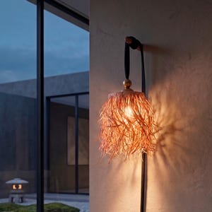 May include: A wall-mounted light fixture featuring a textured, fringed lampshade in a warm tan hue. The shade is suspended by a dark strap attached to a circular wall mount, with a small gold-toned sphere accent connecting the strap to the shade.