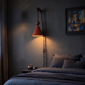 Può includere: Una lampada da parete con un paralume conico color ruggine. La lampada è appesa a un cavo intrecciato e presenta una sfera di legno e un interruttore. La luce calda illumina una parete grigio scuro, con un letto e un'opera d'arte sullo sfondo.