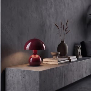 May include: A glossy dark red mushroom-shaped table lamp with a rounded dome shade and a spherical base. The lamp emits a soft light from beneath the shade, highlighting its smooth, reflective surface and distinct curved silhouette.