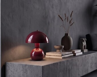 Peut inclure: Une lampe de table rouge foncé brillante en forme de champignon avec un abat-jour en dôme arrondi et une base sphérique. La lampe émet une lumière douce sous l'abat-jour, soulignant sa surface lisse et réfléchissante et sa silhouette courbe.
