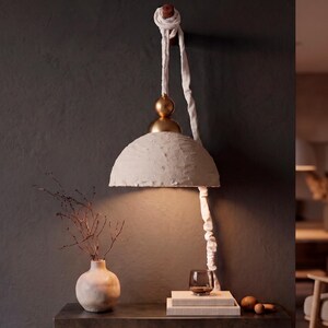 May include: A wall-mounted pendant lamp featuring a textured off-white dome shade, a gold-toned spherical accent, and a white fabric-covered cord that hangs from a wooden peg. The lamp emits a warm light downward, illuminating the surface below.