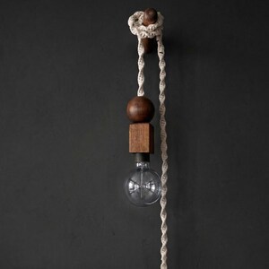 Peut inclure: Un luminaire suspendu moderne avec une sph&egrave;re et un cube en bois, une ampoule en verre transparent et une corde cr&egrave;me. Le luminaire est mont&eacute; sur un mur gris fonc&eacute;. L'ampoule est visible.