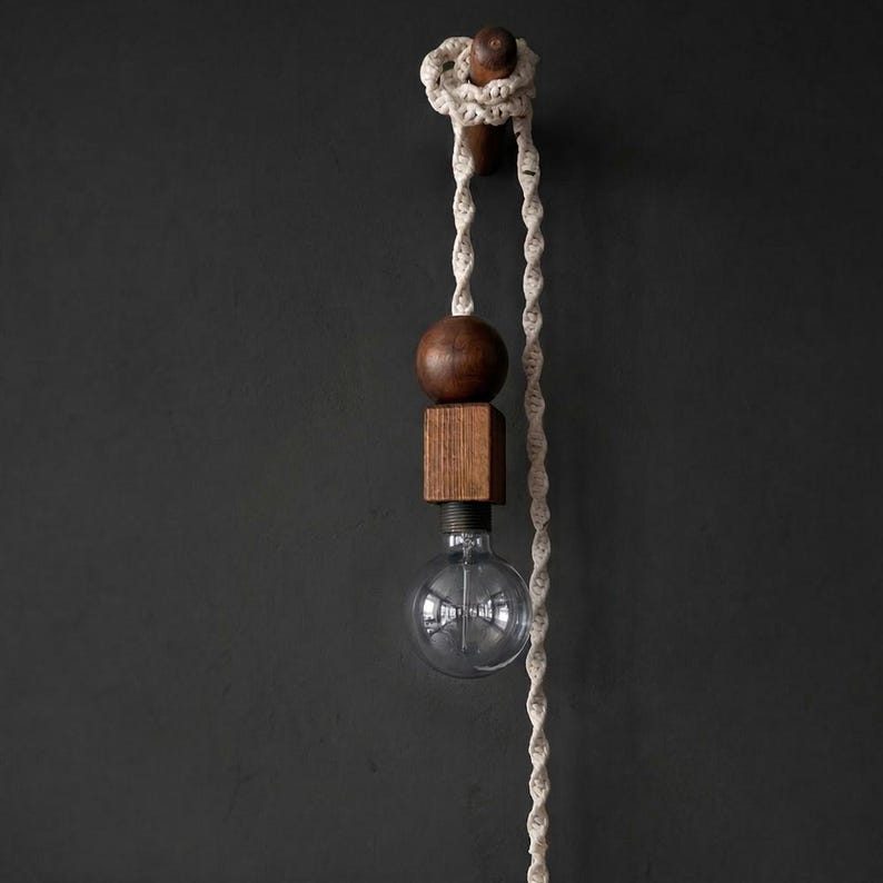 Peut inclure: Un luminaire suspendu moderne avec une sph&egrave;re et un cube en bois, une ampoule en verre transparent et une corde cr&egrave;me. Le luminaire est mont&eacute; sur un mur gris fonc&eacute;. L'ampoule est visible.