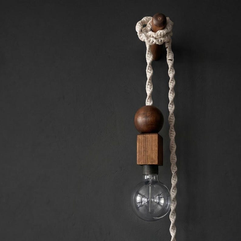 Peut inclure: Suspension luminaire moderne avec une ampoule en verre transparent, une sph&egrave;re et un bloc en bois. La lumi&egrave;re est suspendue &agrave; une corde blanche et fix&eacute;e &agrave; un mur gris fonc&eacute;. Les &eacute;l&eacute;ments en bois ont une finition naturelle et chaleureuse.