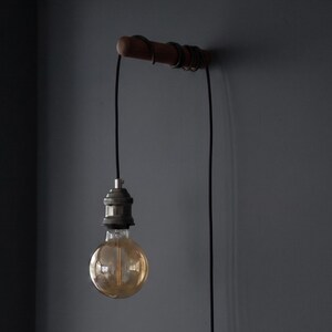 May include: A minimalist wall-mounted light fixture. It features a wooden bar with a coiled wire detail, a black cord, and a vintage-style Edison bulb. The bulb is round and emits a warm, amber glow. The fixture is mounted on a dark gray wall.
