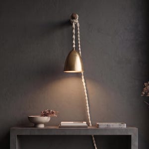 May include: A brass dome-shaped pendant light fixture suspended from a dark wooden wall hook by a long, braided off-white cord. The light emits a warm glow downward, illuminating the space below the fixture with a soft, focused light beam.
