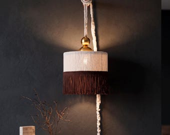 Peut inclure: Une applique murale composée d'un abat-jour cylindrique avec des fils verticaux blancs sur la partie supérieure et des franges marron foncé sur la partie inférieure. Un élément doré sphérique surmonte l'abat-jour, suspendu par un cordon blanc.