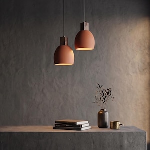 May include: Two terracotta-coloured dome pendant lights with dark brown wooden tops hang from thin cords. Below, a stack of three books, a dark vase containing a thin leafy branch, and a small metallic cup sit on a flat surface.