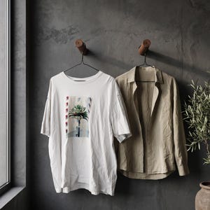 May include: A white short-sleeve t-shirt with a graphic print of a palm tree and the text "back and forth" alongside a beige long-sleeve button-down shirt. Both garments are hanging on black wire hangers from wooden wall hooks.