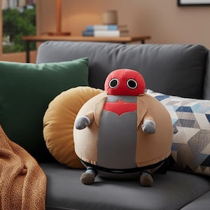 May include: A plush toy of a character with a red helmet, gray shirt, and tan jacket, sitting on a gray couch with pillows and a blanket. The toy has a round body and small arms and legs. The background includes a window, lamp, and side table.