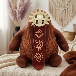 May include: A plush toy in shades of brown, featuring a sun-like headpiece with glasses and a decorative burgundy scarf. The toy has a patterned design on its body and arms, and is sitting on a cream-colored blanket. The overall theme is a whimsical, decorative item.