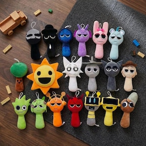 May include: A collection of colorful plush toys with various character designs. The toys include a sun, a green apple, and several figures with unique faces and features. The plushies are arranged on a dark gray surface.