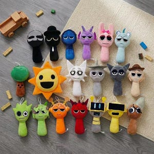 May include: A collection of twenty small plush toys featuring various characters with distinct shapes, colors, and facial expressions, including a yellow sun, a green tree, a red devil, and several humanoid figures, arranged on a textured surface.