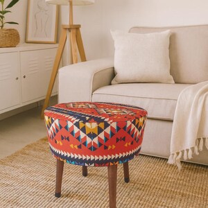 May include: A round upholstered ottoman with a vibrant geometric pattern in red, blue, yellow, and white. The ottoman has dark brown wooden legs and is placed on a woven rug. Home decor.