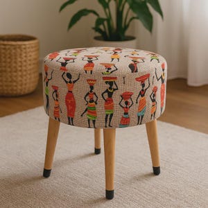 May include: A round, decorative stool with a beige fabric top featuring a repeating pattern of figures carrying items on their heads. The figures are in various colors, including red, orange, green, and black. The stool has four wooden legs.