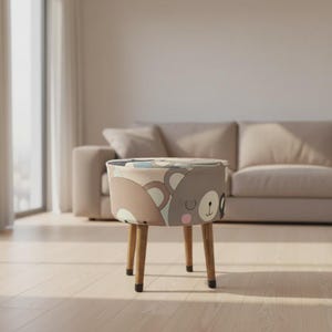 Puede incluir: Un otomán redondo tapizado con un diseño de oso en tonos marrones, grises y azules. El otomán tiene cuatro patas de madera. El fondo incluye un sofá beige y una ventana con una cortina transparente. Aproximadamente 40 cm de diámetro.