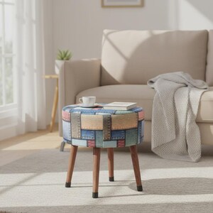 Puede incluir: Un pequeño taburete redondo con un asiento de diseño patchwork en tonos azules, rosas y beige. El taburete tiene cuatro patas de madera y está colocado sobre una alfombra de color claro. Una taza y un libro descansan sobre el taburete.