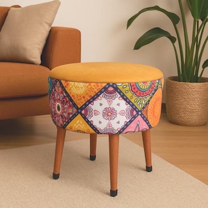 Op de afbeelding: Een ronde, decoratieve kruk met een mosterdgeel kussen en een patroonrok. De rok heeft kleurrijke geometrische ontwerpen. De kruk heeft vier houten poten met zwarte uiteinden. De kruk is ongeveer 40 cm hoog.