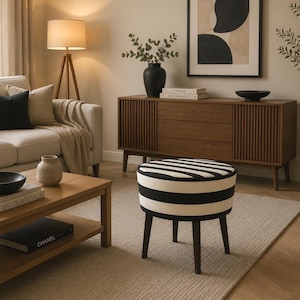 May include: A living room scene featuring a beige sofa with black and beige pillows, a wooden coffee table, and a striped ottoman. A wooden cabinet and a floor lamp are also visible, creating a cozy and inviting atmosphere.