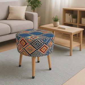 Può includere: Un pouf rotondo con un motivo geometrico blu, arancione e nero. Il pouf ha gambe in legno chiaro con punte nere. Il design ricorda un motivo tribale, adatto all'arredamento della casa.