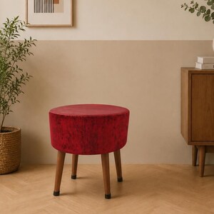 Op de afbeelding: Een ronde, rode fluwelen kruk met houten poten. De kruk is een levendig accentstuk, perfect om een vleugje kleur aan elke kamer toe te voegen. De poten zijn warmbruin, tegen een neutrale achtergrond.