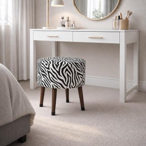 Könnte beinhalten: Ein weißer Schminktisch mit zwei Schubladen und goldenen Griffen, ein runder Spiegel und ein Hocker mit Zebra-Print und dunkelbraunen Beinen. Der Raum hat eine neutrale Farbpalette mit weichem Teppich und weißen Vorhängen.