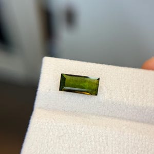 Op de afbeelding: Een rechthoekige, gefacetteerde edelsteen met een diepe olijfgroene tint. De steen heeft een heldere, transparante kwaliteit met zichtbare interne facetten en een gepolijst oppervlak, wat een duidelijke langwerpige vorm laat zien.