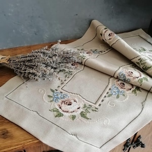 Puede incluir: Camino de mesa de lino beige con diseño floral bordado con rosas rosas y detalles azules. Un manojo de lavanda seca descansa sobre una superficie de madera, complementando la delicada estética del camino de mesa. El camino de mesa está doblado, mostrando el intrincado bordado.