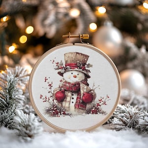 May include: A cross-stitch embroidery of a snowman wearing a top hat, scarf, and mittens. The snowman has a carrot nose and is surrounded by red berries and greenery. The embroidery is set in a wooden hoop, with a snowy background and Christmas tree.