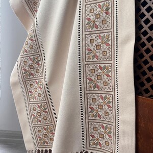 May include: Cream-colored textile with embroidered floral patterns in red, green, and yellow. The design is framed by a brown border and a fringe at the bottom. The fabric is draped over a wooden surface.