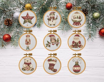 May include: Nine Christmas-themed cross-stitch embroidery hoops. Designs include a star, snowman, snow globe, nutcracker, owl, Santa Claus, teddy bear, stocking, and a vase. The hoops are displayed on a white wooden surface with evergreen branches and ornaments.