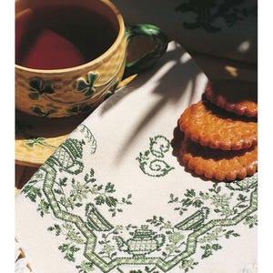 Könnte beinhalten: Eine gelbe Teetasse mit grünem Henkel und Kleeblattmuster. Ein weißes Tuch mit grüner Kreuzstichstickerei mit Teekannen, Vögeln und floralen Mustern. Ein Stapel Kekse liegt neben dem Tuch. Die Szene hat eine Vintage-Atmosphäre.