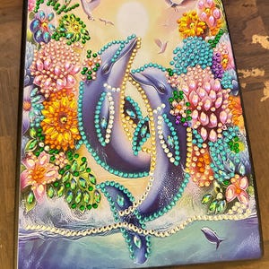 May include: A framed artwork depicting two dolphins, adorned with sparkling embellishments, amidst colourful floral elements. The image includes the ocean, birds, and a setting sun, creating a vibrant and detailed scene. The artwork is framed in black.