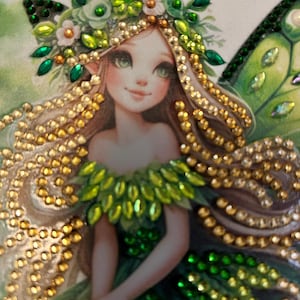 May include: A detailed illustration of a fairy with green eyes, adorned with a floral crown and embellished with gold and green gems. The fairy has long, flowing hair and wears a green dress. The image is a close-up, highlighting the intricate details of the artwork.