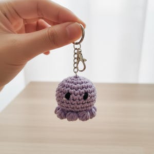 Könnte beinhalten: Ein lavendelfarbener Häkel-Oktopus-Schlüsselanhänger mit schwarzen Augen und acht kleinen Tentakeln. Der Schlüsselanhänger hat einen silberfarbenen Schlüsselring und einen Karabinerverschluss. Der Oktopus schwebt in der Luft.