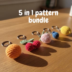 Pode incluir: Uma coleção de cinco chaveiros de crochê em forma de frutas: uma laranja, cerejas, um morango, um mirtilo e um limão. Cada chaveiro tem um anel prateado. O texto "5 in 1 pattern bundle" é exibido acima dos chaveiros.