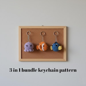Può includere: Tre portachiavi all'uncinetto: un polpo viola, un pesce arancione e bianco e un pesce blu con pinne gialle. Sono appesi a una bacheca di sughero in una cornice di legno. Il testo "3 in 1 bundle keychain pattern" è sotto la cornice.