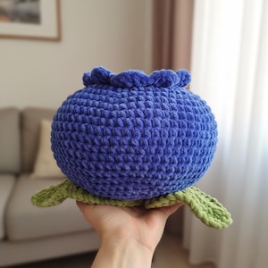 Może przedstawiać: Ręcznie robiona zabawka w kształcie jagody, wykonana na szydełku. Okrągły, teksturowany korpus jest w kolorze głębokiego niebieskiego, z falbanką na górze i zielonymi liśćmi u podstawy. Zabawka ma przypominać jagodę.