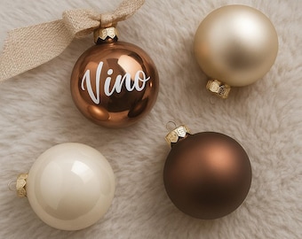 Könnte beinhalten: Vier Weihnachtskugeln in Braun- und Cremetönen liegen auf einer weichen, weißen Oberfläche. Eine Kugel ist bronzefarben mit dem Wort "Vino" in weißer Schrift. Die anderen Kugeln sind cremefarben, goldfarben und dunkelbraun.